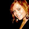 A static image of a person with light brown hair, wearing large circular earrings and a dark top, against a black background.
