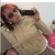 A person in a beige sweater holds a black phone to their ear, seated on a pink and white patterned couch with light-colored walls behind them.