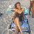 A pixelated image of a woman in a black swimsuit standing on a beach with blue water and sand, holding a light-colored object.