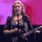 A woman with blonde hair sings into a microphone while holding an electric guitar, set against alternating pink and red backgrounds.