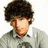 A person with dark curly hair appears in three distinct poses: looking down, facing forward with a red heart symbol, and squinting against green foliage.
