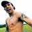 A pixelated image of a shirtless man with dark hair and a beard, standing outdoors with a green field and blue sky background.