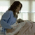A pixelated figure in a blue top and light pants sits at a table, facing right with long dark hair, against a bright window background.
