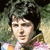 Pixelated image of a person with dark hair and light skin, wearing a colorful garment against a blurred green and brown background.