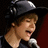 Pixelated image of a person wearing a dark hat and holding a microphone, with a dark background and red vertical element.