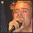 A person with light brown hair holds a microphone near their mouth, eyes closed in a static image.
