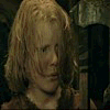 A young person with light brown hair in a dark setting, holding an axe-like object while turning their head