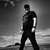 A grayscale pixelated figure in a dark outfit stands against a gradient background with light and dark tones.