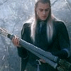 A person with long light hair holds a large sword-like object, standing in front of blurred trees while text appears below.