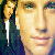 Pixelated close-up of two figures with light skin tones, one in dark clothing with a white stripe, the other in a light-colored top, against a blurred background.