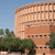A pixelated image of a large cylindrical building with vertical lines and rectangular windows, set against a clear blue sky with green trees in the foreground.