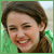 A pixelated close-up of a person with brown hair, smiling broadly with visible teeth and closed eyes against a green background.