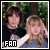 Two people pose together outdoors with green foliage background; one wears dark clothing, the other light-colored top and blue jeans; text 'FAN' appears in pixelated white letters on black rectangle at bottom left.