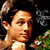 A pixelated image of a person with dark hair and light skin, wearing no shirt, against a background with green foliage and red flowers.