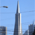 A static pixelated image shows a tall silver skyscraper with a pointed spire against a light blue sky, with smaller buildings visible on the left and right edges.