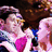 Pixelated image of two figures in close physical contact, one with light hair and pink clothing against a dark background.