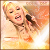 A woman with blonde hair sings into a black microphone, wearing a light-colored top with orange accents, against a warm-toned background with 'COME ON!' text.