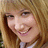 Pixelated close-up of a smiling person with light brown hair and visible teeth, wearing a white top against a blurred background.