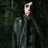 Pixelated figure in dark green coat with light-colored collar, standing against dark green foliage background, face obscured by pixelation.