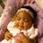 Pixelated image of a child with closed eyes wearing a white ruffled collar and brown outfit, held by an adult in pink-toned clothing.