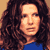 Pixelated static image of a person with long dark curly hair wearing a blue top against a light brown background.