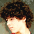 A person with curly brown hair in profile view, wearing a red top against a muted green background.