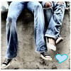 Two people in blue jeans and sneakers stand on a textured surface with a small light blue heart symbol in the bottom right corner.