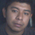 Pixelated close-up of a person's face with neutral expression, visible ear and partial neck against dark background.