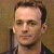 Pixelated portrait of a person with short dark hair, light skin, and a neutral expression, wearing a dark jacket over a light shirt against a brownish background.