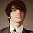 A static image of a person in a brown suit with a white shirt and black tie, holding a guitar, with orange text reading "Drake Bell" below.