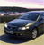 A dark blue sedan with headlights on, positioned on a light-colored road under a gradient sky, rendered in low-resolution pixel art style.