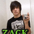 A person in a black and white graphic t-shirt holds a silver digital camera, with green text 'ZACK' overlaid on the bottom-left frame.