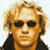 Pixelated static image of a person with light brown curly hair, dark sunglasses, and black clothing against a white background.