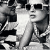 Pixelated grayscale image of a person wearing dark sunglasses and light-colored clothing, standing against a patterned background.