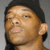 Two individuals with dark skin tones appear side-by-side against a plain gray background, one wearing a black beanie and the other a camouflage-patterned cap.