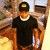 A person wearing a black cap with a yellow rectangle, black shirt, white collar, and brown pants stands against a blurred background with light and dark tones.
