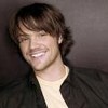 Static close-up of a person with brown hair wearing a dark jacket over a light shirt against a gray background.