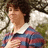 Pixelated image of a person with short dark hair wearing a red and blue striped shirt over a white collar, standing outdoors near greenery.