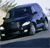 Pixelated image of a dark blue sedan with visible headlights and side mirrors, positioned on a road with blurred background elements.