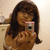 A person with long brown hair holds a silver digital camera with a pink top, taking a mirror selfie in a room with light-colored walls and a cabinet.