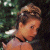 A pixelated image of a person with light brown hair styled up, wearing a black choker and pink top against dark green foliage background.