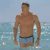 A pixelated static image shows a shirtless man standing in shallow water, wearing blue swim trunks with a dark waistband, against a light blue sky and turquoise sea background.