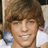 Pixelated close-up of a person with brown hair and light skin, wearing a white shirt, against a blurred background with blue and white elements.