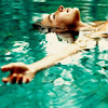 A person floats on their back in turquoise water with arms extended, head tilted back, eyes closed, and mouth slightly open.