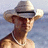 Pixelated image of a person wearing a light-colored cowboy hat and pearl necklace against a blurred background, with blue text on a bright green field.