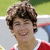 A person with curly brown hair wearing a red and white shirt smiles broadly in a of four with varying background details