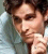 Pixelated close-up of a person with dark curly hair resting their chin on one hand, wearing a light blue shirt against a blurred background.