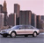 Pixelated cityscape with tall buildings and a silver sedan on a road under a light pink sky.