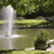 Static pixelated image of a white fountain structure with water spray, set against a green landscape with trees and a body of water.