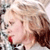 A pixelated close-up of a person with light hair and red lips, wearing a dark top with a light collar, against a blurred background.